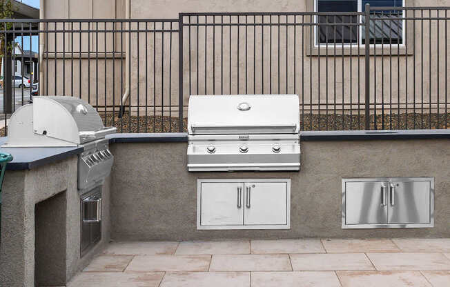 A white grill is on a patio next to a wall and a window.
