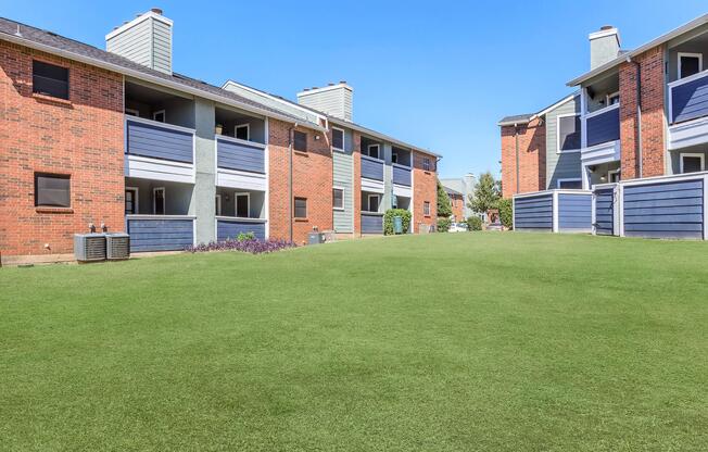 View of a well-maintained apartment complex featuring two buildings with a mix of brick and blue siding. The grassy lawn in the foreground is neatly trimmed, and the overall setting is sunny and inviting. Air conditioning units are visible on the left side of the image.