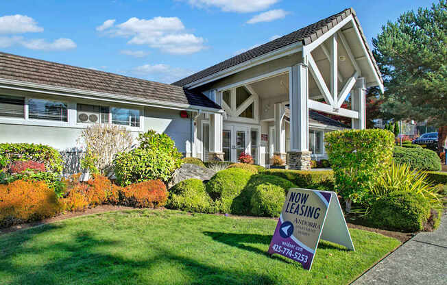 A welcoming leasing office exterior here at Andorra featuring a covered entry with white trim and stone accents, large windows, and neatly landscaped shrubs and greenery, with a sidewalk and lawn in the foreground and a visible now leasing sign, creating an inviting first impression for prospective residents.
