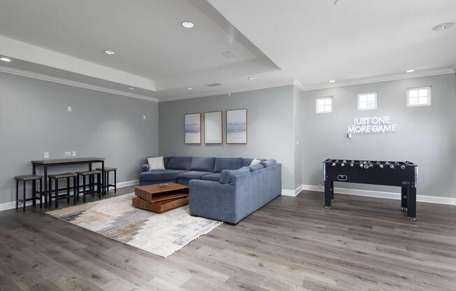 A spacious game room featuring a blue sectional sofa, a wooden coffee table, a foosball table, and a small dining table with chairs. The walls are painted light gray, and three framed pictures are arranged above the sofa. A wall-mounted sign reads "JUST ONE MORE GAME."