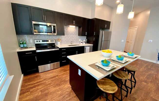 A large kitchen with a breakfast bar, three pendant lights, stainless steel appliances, hardwood-style flooring, a window, and black cabinets.