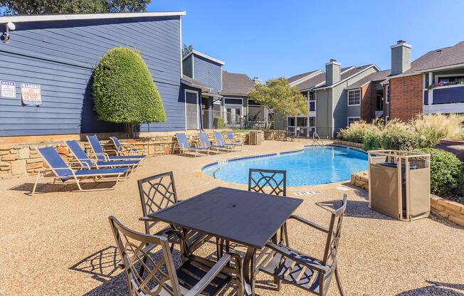 A swimming pool surrounded by lounge chairs and a table with chairs, set in a landscaped area with buildings in the background. The scene is under a clear blue sky, creating a relaxing outdoor atmosphere.