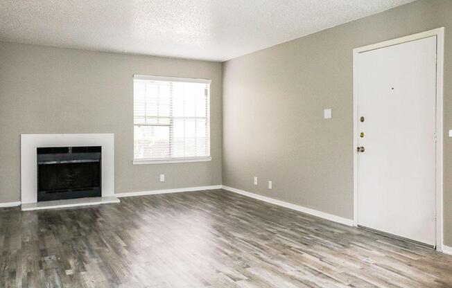 A spacious, unfurnished living room with light gray walls and a large window letting in natural light. The floor is made of light-colored wood-laminate. There's a white fireplace on one wall and a white door leading outside, creating a simple and modern atmosphere.