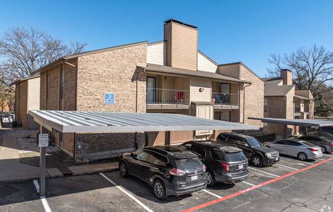 A covered parking area with cars and an apartment building in the background at Preston Oaks Apartments in Dallas, TX.