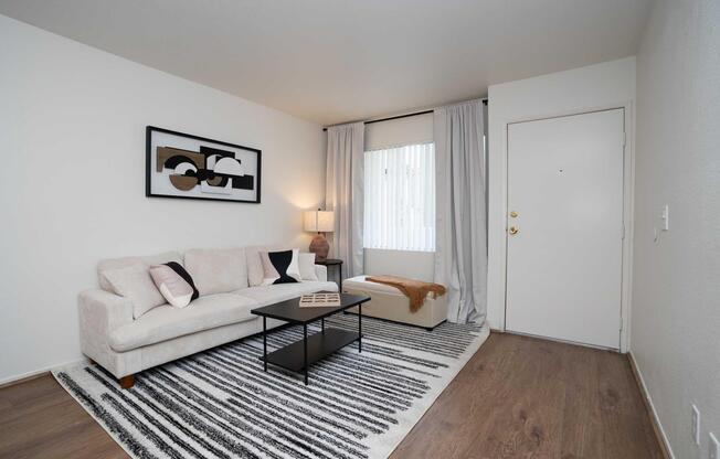 A minimalist living room featuring a light-colored sofa with decorative pillows, a black coffee table on a striped rug, and a warm throw. A floor lamp adds soft lighting, and there are sheer curtains covering a window. The entrance door is visible, and a piece of abstract wall art hangs above the sofa.