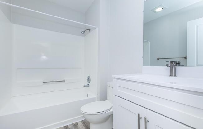 A clean, modern bathroom featuring a white bathtub with a shower, a wall-mounted mirror above a sleek white vanity with a sink, and neutral-colored walls. The flooring is light wood, enhancing the bright and fresh atmosphere of the space.
