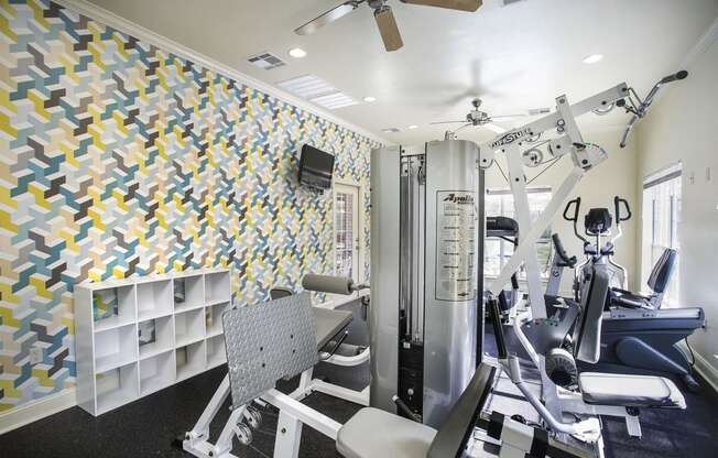 Tyler, TX Apartments - Community Fitness Center with Weight Machines, a Ceiling Fan, and Large Windows.