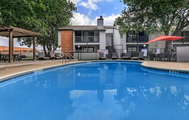 A clear blue swimming pool surrounded by lounge chairs and an umbrella, with apartment buildings in the background and trees providing shade.
