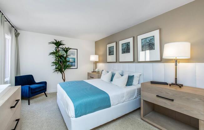A modern bedroom featuring a bed with a light blue throw blanket, flanked by nightstands with lamps. There's a blue accent chair in the corner, a potted plant, and wall art depicting abstract designs. Large windows allow natural light to fill the room, creating a calm and inviting atmosphere.