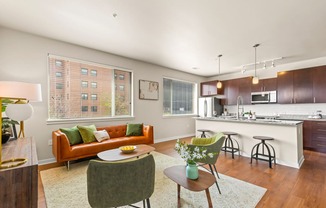 Modern living room with an orange couch, green accent chairs and oversized windows, complete with view into kitchen with barstool seating and pendant lighting at Sylvan Uptown, Denver, CO, 80203