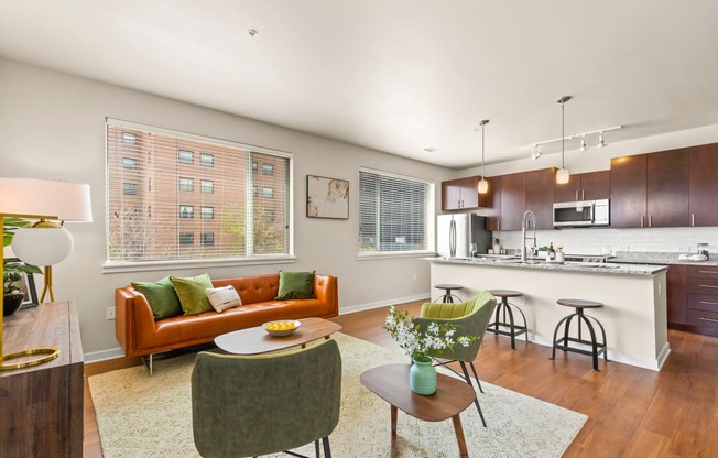 Modern living room with an orange couch, green accent chairs and oversized windows, complete with view into kitchen with barstool seating and pendant lighting at Sylvan Uptown, Denver, CO, 80203