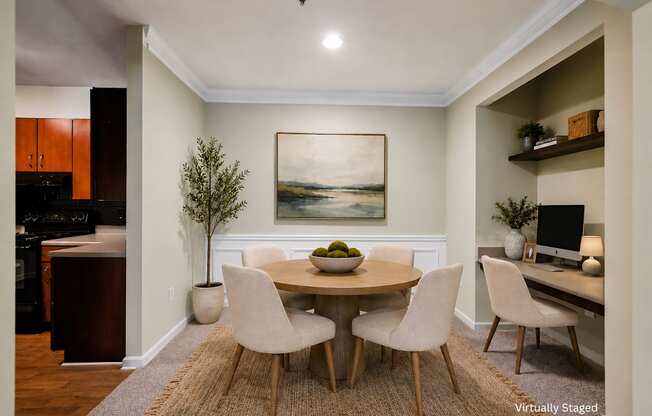 A dining room with a round table and chairs, a painting on the wall, and a kitchen area in the background.