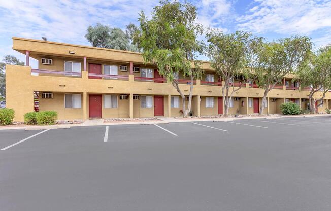 A view of a two-story motel with tan walls and red doors, surrounded by trees. The parking lot in front is empty, and the sky is partly cloudy. The building features multiple rooms with a balcony style, creating a simple and functional design.