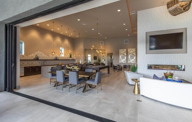 A spacious modern living area featuring a large open layout with stylish furniture. There are grey upholstered chairs and tables, a cozy fireplace, and elegant lighting fixtures. The walls are a light color with decorative accents, and large windows provide a view of the outdoor area.