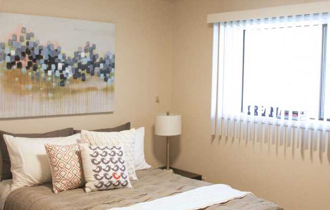 Bedroom with queen sized bed and large window with blinds. Staged with bed, side table, lamp and wall art.at The Lakes Apartments, Moses Lake, 98837