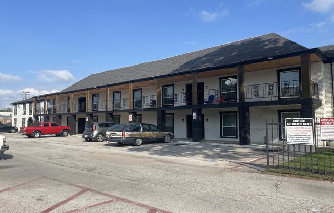 Two-story building with multiple balconies, each with chairs. The ground level has parked cars, including a red pickup truck and a gray sedan. A sign indicates caution for automatic gates. The building is set against a blue sky with some clouds, and there are a few trees in the background.