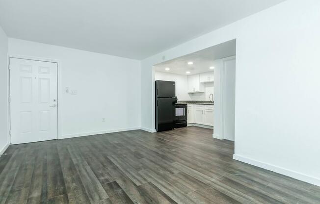 A spacious, modern apartment interior featuring an open layout. The room has light-colored walls and dark wooden flooring. To the left, there's a white door leading outside. In the background, a kitchen area with sleek cabinets and a black refrigerator is visible, illuminated by recessed lighting.