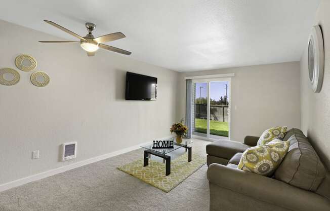 A Furnished Living Room with Carpeting, Sliding Glass Doors Leading to Outdoor Area, and Overhead Ceiling Fan with Lighting at Campo Basso Apartment Homes, Lynnwood, WA