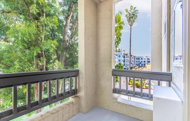 A balcony with a view of lush green trees and a palm tree, overlooking an apartment complex. The space has a light-colored wall and railing, and the floor is concrete. Natural light is streaming in, creating a bright and inviting atmosphere.