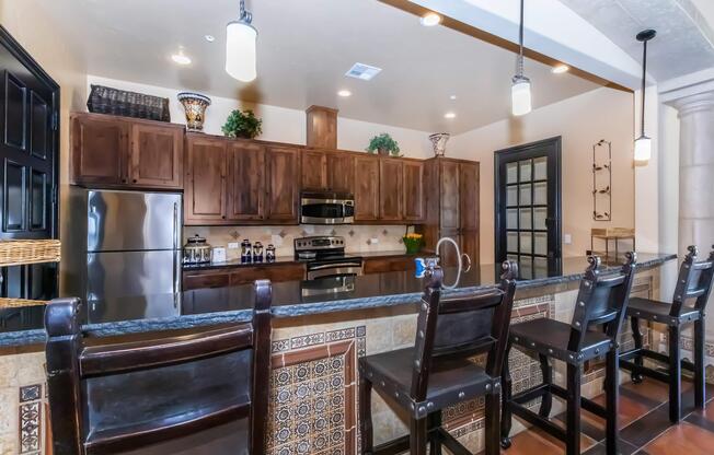 A modern kitchen featuring dark wooden cabinets and stainless steel appliances. The countertop has decorative tiles, and there are several barstools lined up against it. Pendant lights hang from the ceiling, adding warmth to the space. The kitchen includes decorative plants and a stylish backsplash.