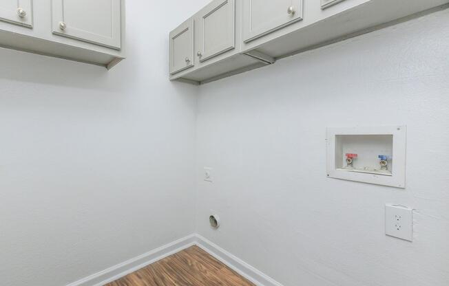 A clean, empty laundry room with light gray cabinets mounted on the wall. The wall is painted white, and there are plumbing connections visible, including hot and cold water valves. The floor features light wood-like laminate, contributing to a modern and tidy appearance.