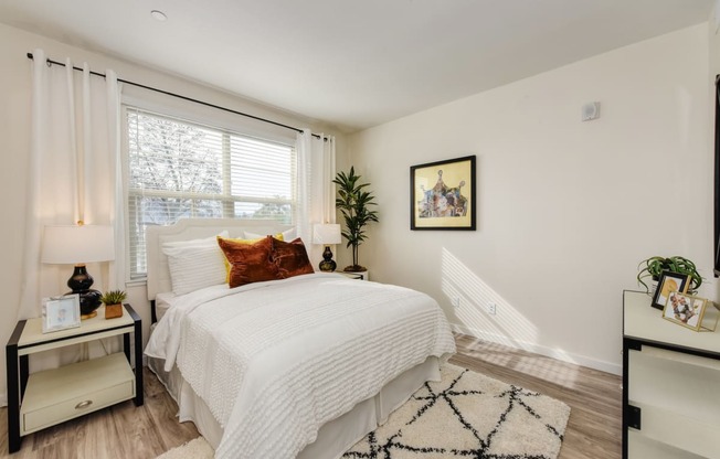 Master bedroom with queen size bed under the large window. Nightstand table with lamp on the side of the bed. Faux plant in corner of the room at Morgan Ranch Apartments, Morgan Hill, California