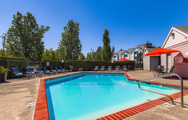 Happy Valley, OR, Apartments - Sunnyside Village - an Outdoor Gated Pool Area with Lounge Seating, Tables with Chairs and Umbrellas, Surrounding Trees and Shrubbery, and a View of the Property Building