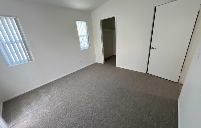 A simple, empty room with beige carpet and light-colored walls. It features two small windows on the left side and a closet with a door on the right. The room has a sloped ceiling, creating a spacious feel despite its minimal furnishings. Natural light enters through the windows, enhancing the bright atmosphere.
