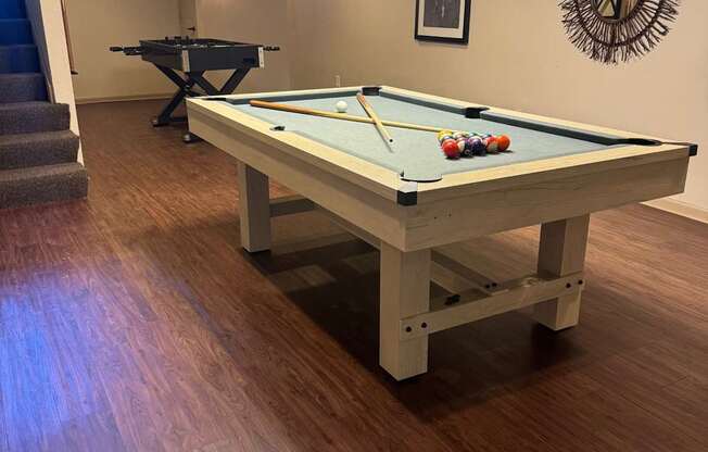 A pool table with a green cloth and white cues is in the middle of a room with a wooden floor and a mirror on the wall.