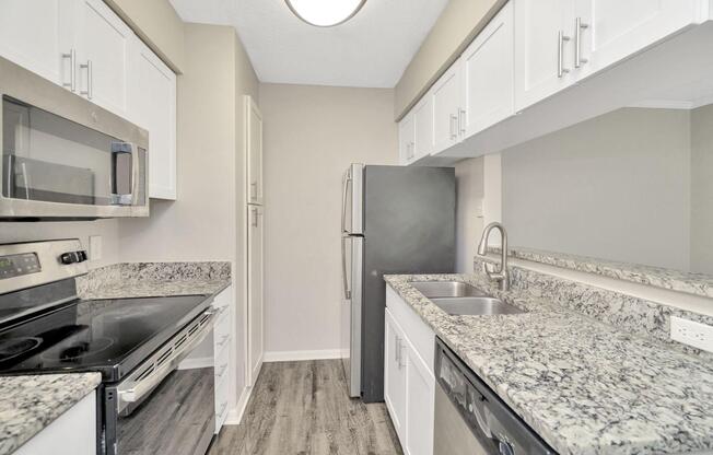 Modern kitchen featuring white cabinetry, granite countertops, a stainless steel microwave, oven, and dishwasher. A black refrigerator is positioned next to the sink, with light-colored walls and a laminate floor enhancing the bright, inviting atmosphere. A ceiling light fixture provides illumination.