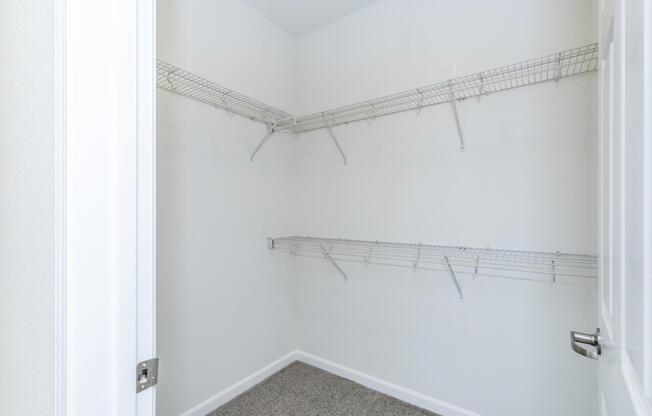 A clean, empty closet featuring white wire shelving on the walls. The walls are a light color, and the floor is covered with carpet. The closet has a simple design, with no items stored inside, creating a spacious and organized appearance.