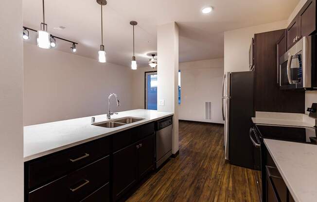 A modern kitchen here at The Park featuring dark wood cabinets, a white island with sink, stainless steel appliances, pendant lighting, and warm wood floors, creating a bright and stylish cooking space.
