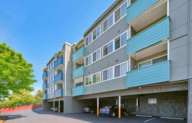 Blue, teal, and beige apartment building with carports underneath. Green trees line the left side of the paved driveway.