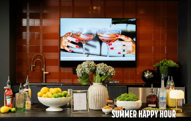 A kitchen counter with a bowl of lemons, a vase with flowers, and a bottle of alcohol with a TV screen showing a cocktail in the background.
