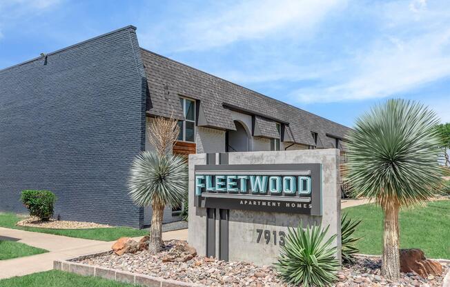 Signage for Fleetwood Apartment Homes in front of a modern building. The structure features a dark exterior with a distinctive roofline and landscaped greenery, including desert plants and rocks. The setting is sunny with a clear blue sky and well-maintained lawn.