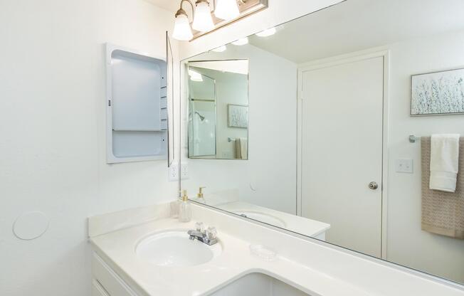 A clean and modern bathroom with a white countertop, sink, and mirror. The walls are painted light, and there are light fixtures above the mirror. A towel hangs on the wall and a decorative framed piece is visible. A medication cabinet is mounted beside the mirror. Overall, the space appears bright and organized.