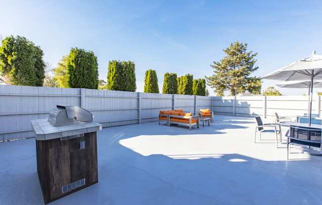 Outdoor lounge with umbrella-covered tables, gas BBQ, and an oversized orange patio couch and coffee table.at The Lakes Apartments, Moses Lake, WA