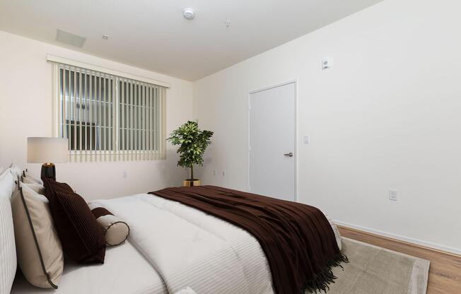 A cozy, modern bedroom featuring a neatly made bed with white linens and a brown throw blanket. A decorative lamp sits on a nightstand beside the bed. Thereâs a tall green plant in the corner and a door leading to another room. The walls are painted light, creating a bright and inviting atmosphere.