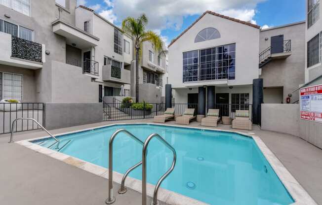 Community Swimming pool located in the middle of the apartment community with apartment buildings overlooking the pool deck. There are wicker lounge chairs present.