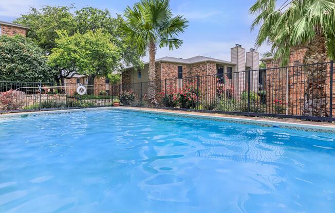 A clear blue swimming pool surrounded by lush greenery and flowering bushes, with a fence in the background and residential buildings visible.