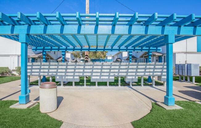 A blue pergola with a grid pattern stands over a row of mailboxes in a residential area. There is a circular trash can nearby, and well-manicured grass surrounds the area. In the background, there are white buildings and clear blue skies.