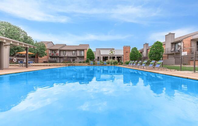 Swimming pool surrounded by lounge chairs, with green trees and apartment buildings in the background. The water is clear and reflects the blue sky. A shaded area with seating is visible to the left.