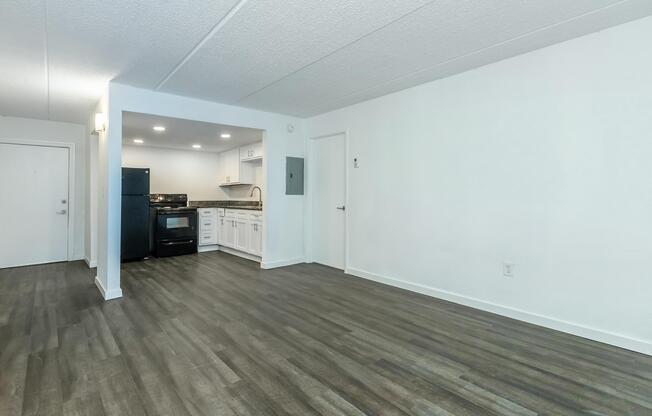 A bright, spacious living area with light gray walls and dark wood-like flooring. On the left, there's a doorway leading outside. The kitchen is partially visible, featuring white cabinets and black appliances. The overall space is clean and modern, offering a welcoming atmosphere.