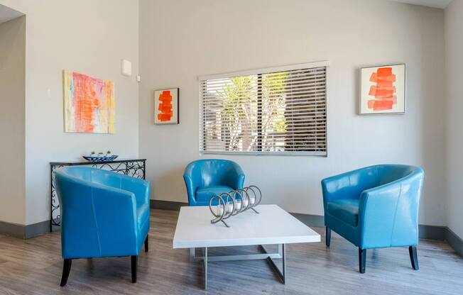 Three blue chairs are arranged around a white coffee table in a room with two paintings on the wall.