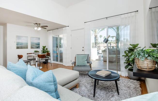 Bright and airy living space featuring a light-colored sofa with blue accent pillows, a round black coffee table, and a plush area rug. Large sliding glass doors open to an outdoor patio, and there are plants and seating in the background. Modern dining area with a table and chairs visible in the distance.