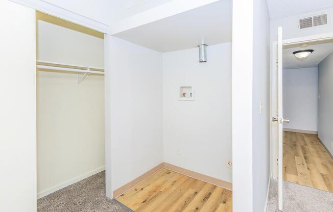 A small, empty room with light-colored walls and wooden flooring. To the left, there is an open closet with a hanging rod. On the right, a doorway leads to another area, and there is a ceiling light visible. The space appears clean and uncluttered, suitable for storage or as a small office.