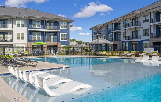 Luxury Apartments for Rent in Huntsville AL - The Kelvin - Gated Sparkling Pool with Sundeck, Submerged and Poolside Lounge Seats, Umbrellas, and View of Building Exterior in Background