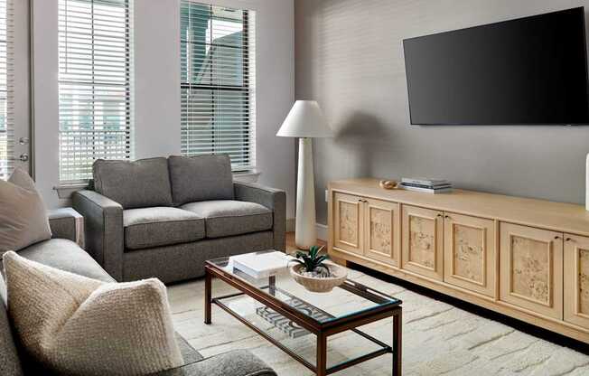 A living room with a grey couch, a coffee table, a lamp, and a television mounted on the wall.