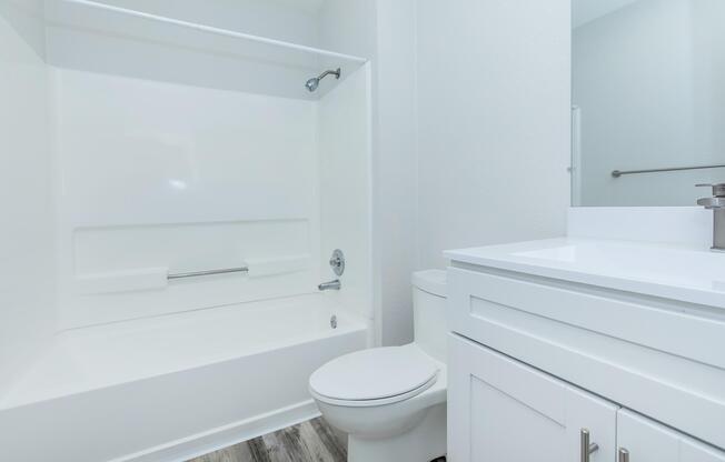 A clean, modern bathroom featuring a white bathtub with a showerhead, a white toilet, and a sleek vanity with a sink. The walls are painted light, and the flooring is a wood-like material, creating a bright and airy atmosphere.