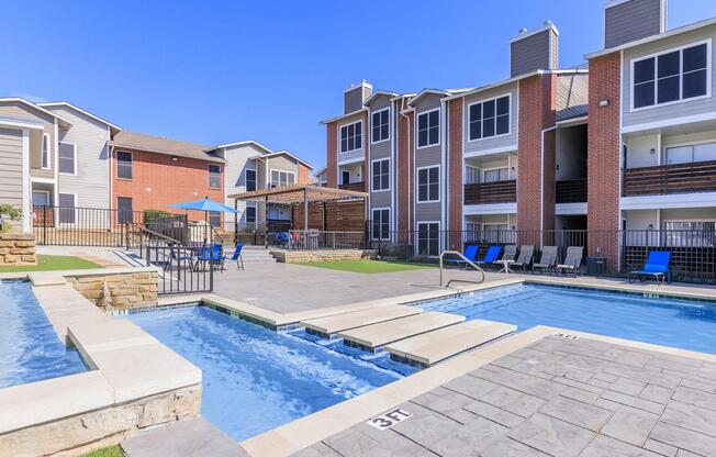 A well-designed outdoor pool area featuring two pools, one with a spa-like section. Surrounding the pools are lounge chairs and tables, with two apartment buildings in the background. The sky is clear and blue, creating a warm and inviting atmosphere.